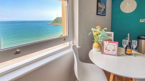 A dining area with a view of the sea, a table with a bottle of champagne, glasses, a vase with flowers, and a framed picture on the wall. - 2 bedroom accommodation in Scarborough (Scarborough)