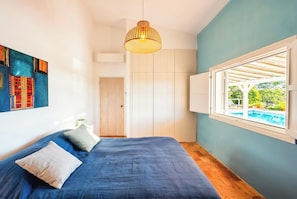 Ein Schlafzimmer mit einem Bett, einer Hängeleuchte, einem Fenster mit Blick auf einen Pool und einer Tür zu einem anderen Raum.