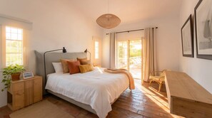 A bedroom with a bed, bedside table, lamp, potted plant, and a view of the outdoors.