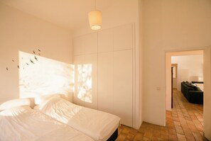 Quarto com cama, luminária pendente e vista para um corredor.
