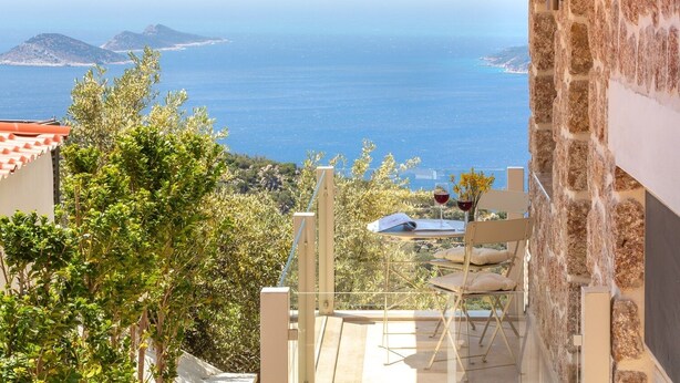 A terrace with a table, two chairs, and a glass of red wine, overlooking a coastal view with the sea and distant islands.