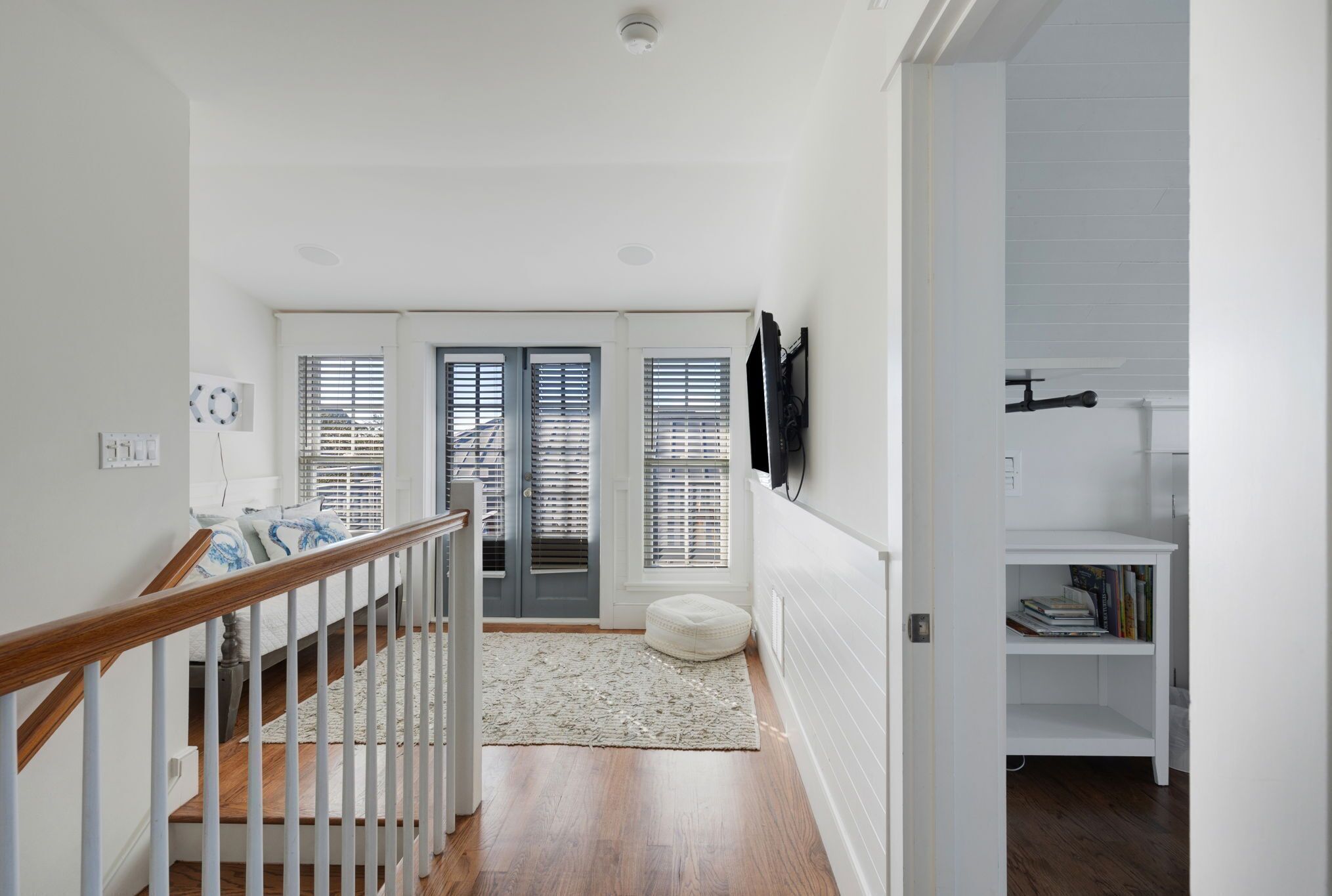A hallway with a wooden staircase, a white door with blinds, and a white shelf with books.