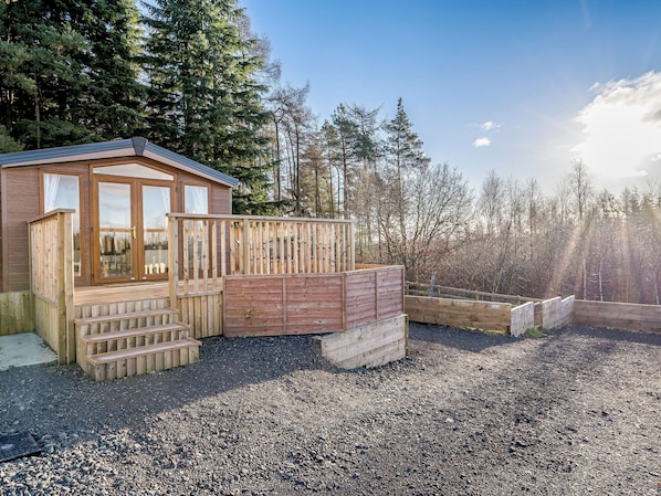 A wooden cabin with a deck, surrounded by a gravel area and dense forest. - The Paddocks - UK7112 (Dollar)