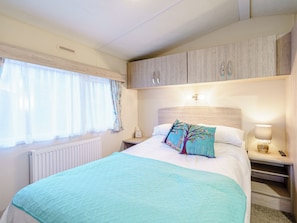 A bedroom with a bed, a nightstand, a lamp, a window with curtains, and a radiator. - The Paddocks - UK7112 (Dollar)