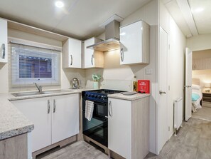 A compact kitchen with white cabinets, a stove, a sink, and a window with blinds. - The Paddocks - UK7112 (Dollar)