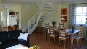 A living room with a black leather sofa, wooden dining table with chairs, a staircase, and a dining area with a window.