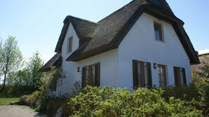 A white house with a steeply pitched roof and green shutters, surrounded by greenery.
