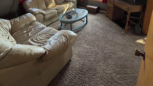 Una sala de estar con ventilador de techo, dos sofás de cuero, una mesa de centro, un televisor y un ventanal con cortinas.
