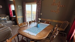 Un comedor con una mesa y sillas de madera, un jarrón azul con una flor blanca, y una pared con un crucigrama.