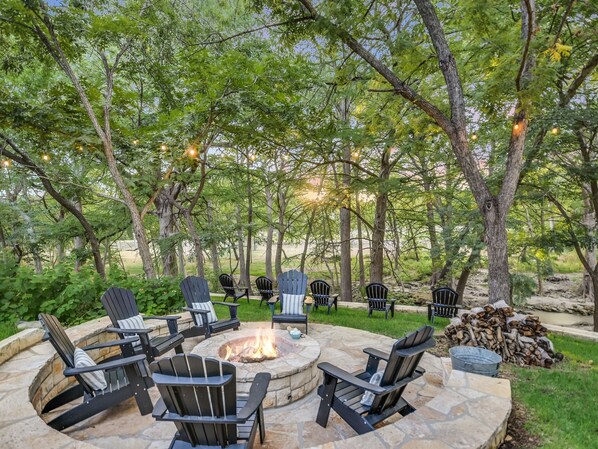 A circular stone fire pit surrounded by Adirondack chairs, set in a wooded outdoor area with trees and string lights.