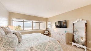 A bedroom with a large bed, a dresser, a TV, a mirror, and a window with a view.