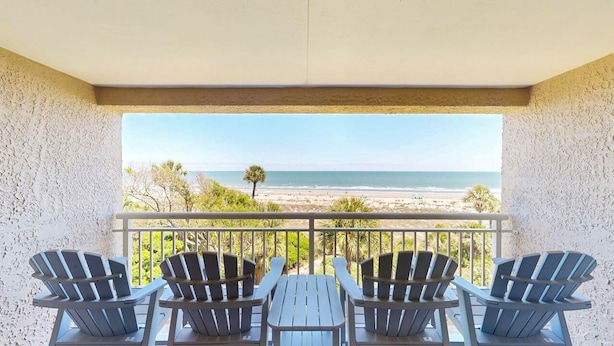 A balcony with four Adirondack chairs and a table, overlooking a beach with a clear view of the ocean and distant trees. - Updated Two Bedroom Fourth Floor Captains Walk Condo, Oceanfront Views (Hilton Head)