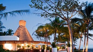 A resort with a thatched-roof building, palm trees, and a view of the ocean.