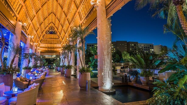 A well-lit outdoor lounge area with a thatched roof, seating arrangements, and a central water feature.