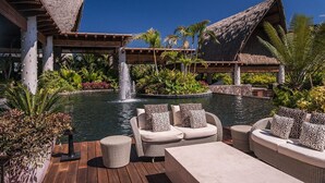 A resort pool area with a waterfall, lounge chairs, and a thatched-roof building.