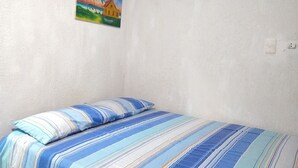 Habitación con cama individual, colcha a rayas, un cuadro de una escena tropical y un equipo de aire acondicionado blanco en la pared.