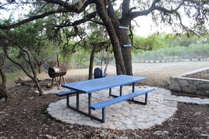 A picnic table with benches, a barbecue grill, and a wooden chair under a large tree. - Concan Frio River Cabin  (Concan)