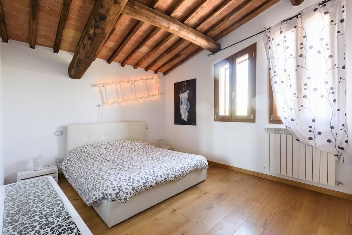 Ein Schlafzimmer mit Bett, Holzbalken an der Decke, einem Heizkörper und einem Fenster mit Vorhängen.