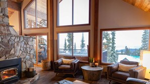 A spacious living room with a stone fireplace, leather sofas, and large windows offering views of trees and a snowy landscape. - Big White Is Calling -  Big White BC (Beaverdell)