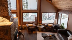 A cozy interior with a stone fireplace, leather sofas, and large windows offering a view of a snowy landscape. - Big White Is Calling -  Big White BC (Beaverdell)