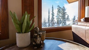 A room with a wooden floor, a window with a snowy forest view, a potted plant, and a small bear figurine on a table. - Big White Is Calling -  Big White BC (Beaverdell)