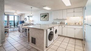A modern kitchen with a central island, built-in washer and dryer, white cabinetry, and a large window overlooking the ocean. - 3 Bedroom/2 Bath Oceanfront Timeshare in Myrtle Beach (Murrells Inlet)