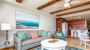 Un salon avec un canapé gris, un fauteuil turquoise et une table basse blanche. On aperçoit, au fond, une cuisine avec des meubles en bois et un réfrigérateur.