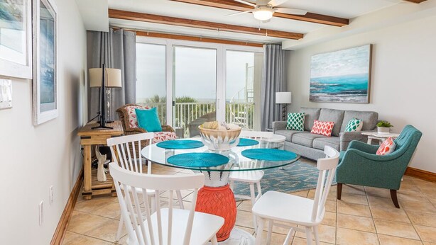 Un espace repas avec une table à plateau de verre entourée de chaises blanches, un plafond en bois et une vue sur l’extérieur grâce à de grandes fenêtres.