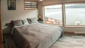 A bedroom with a large bed, a view of a lake, and a painting on the wall. - Lakefront Hideaway w/ beautiful views, quiet area, private lake access & dock! (Christina Lake)