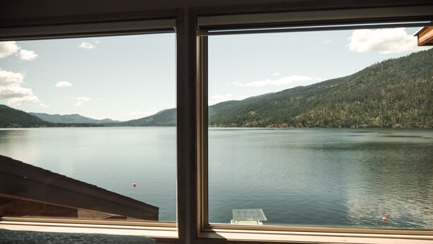 A room with a large window overlooking a calm lake and forested mountains. - Lakefront Hideaway w/ beautiful views, quiet area, private lake access & dock! (Christina Lake)