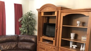 A living room with a brown leather sofa, a wooden cabinet with shelves, and a television. - Brick House Retreat at the Heart of Hamlet (HAMLET)