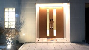 Entrada de um edifício moderno, com porta de madeira, iluminada por luzes no teto, e um caminho de azulejos que a leva até ela.