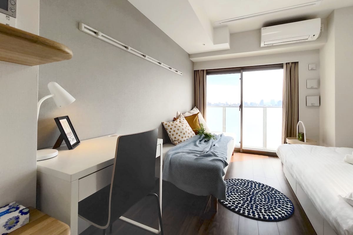 A modern hotel room with a desk, chair, bed, and a view of the city through large windows.