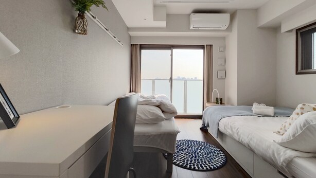 Quarto de hotel moderno com cama, escrivaninha, cadeira, tapete redondo, planta em vaso e vista para a cidade pela janela.