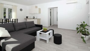 Una sala moderna con un sofá seccional gris, una mesita auxiliar blanca pequeña y un reposapiés negro. Se puede ver, al fondo, un comedor con una mesa y sillas.
