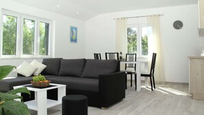 Un moderno salón con un sofá gris, una mesa de centro blanca y un reposapiés negro. Hay una zona de comedor con una mesa blanca y sillas negras. Amplias ventanas dejan entrar luz natural y ofrecen vistas al follaje exterior.