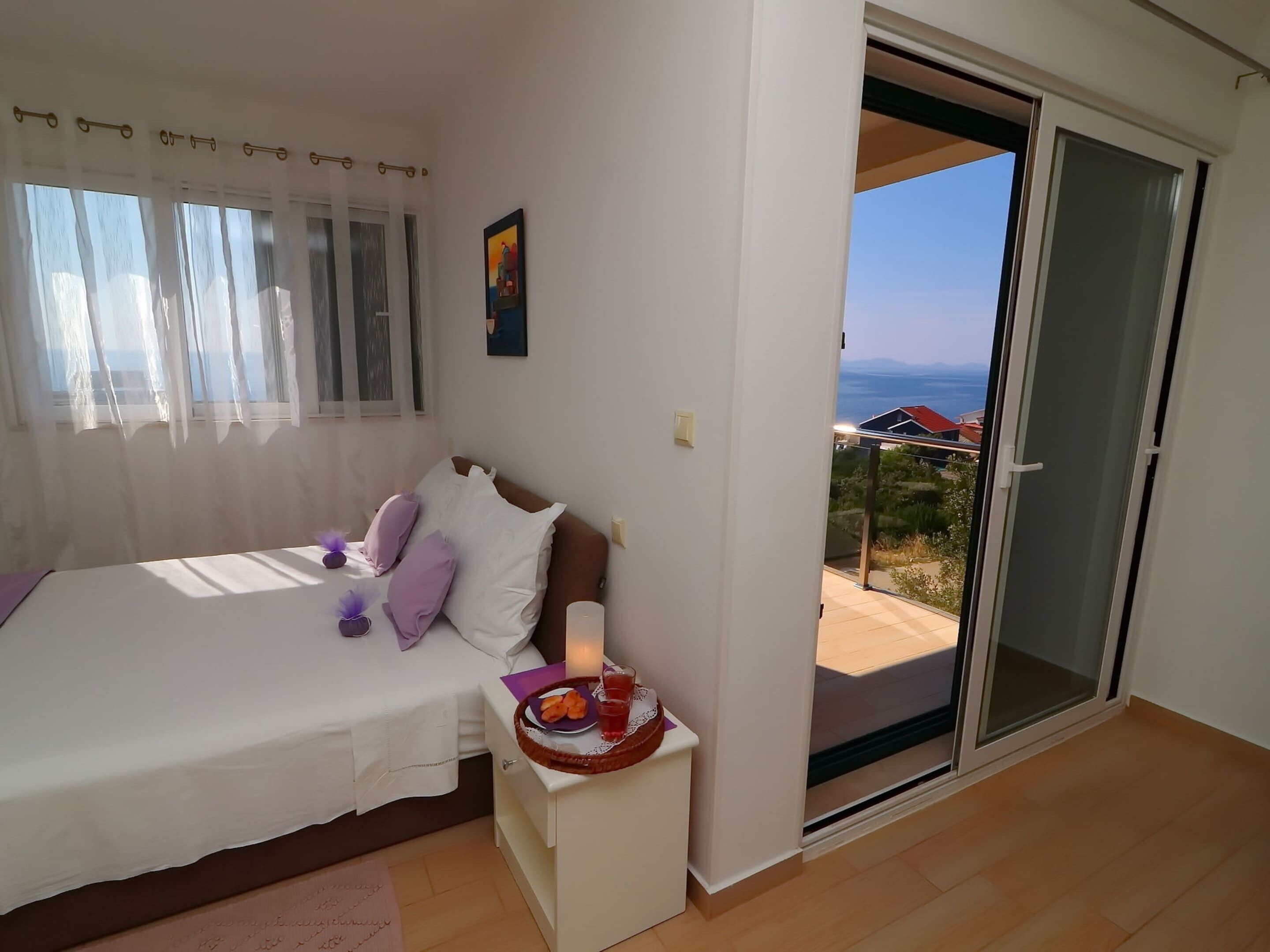 A bedroom with a bed, a nightstand with a tray of breakfast, a painting on the wall, and a view of the outdoors through a sliding door.