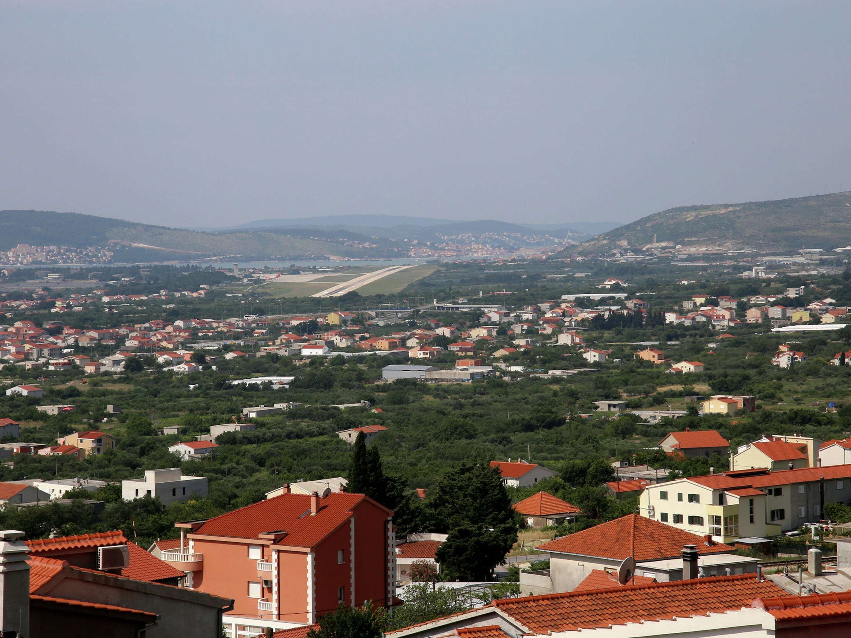 A residential area with red-tiled roofs, a mix of single and multi-story buildings, and a distant view of a coastline.