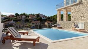 A swimming pool with lounge chairs, a stone building with a balcony, and houses in the background.