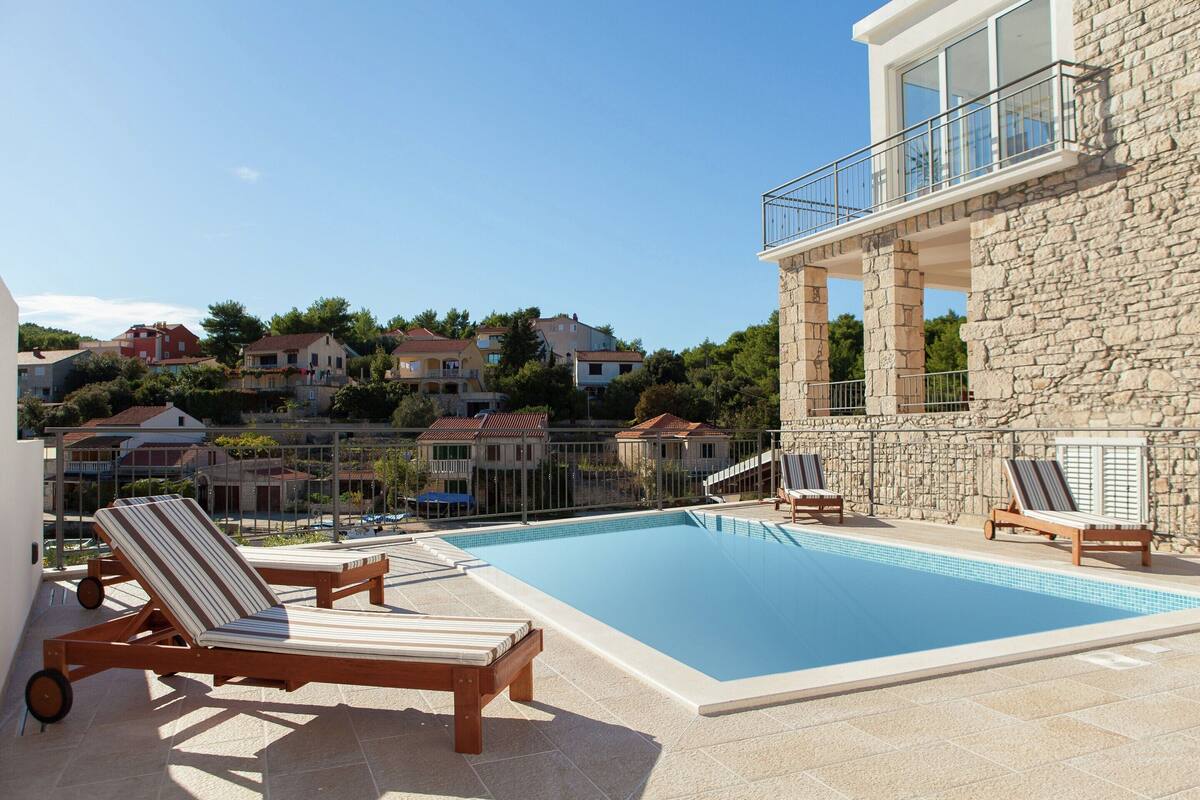Una piscina con sillas de descanso, un edificio de piedra con balcón y casas al fondo.
