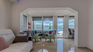 A spacious living area with a sofa, colorful chairs, a table, and a view of the sea and mountains through large windows. - Apartment in Arbanija With Terrace (Trogir)