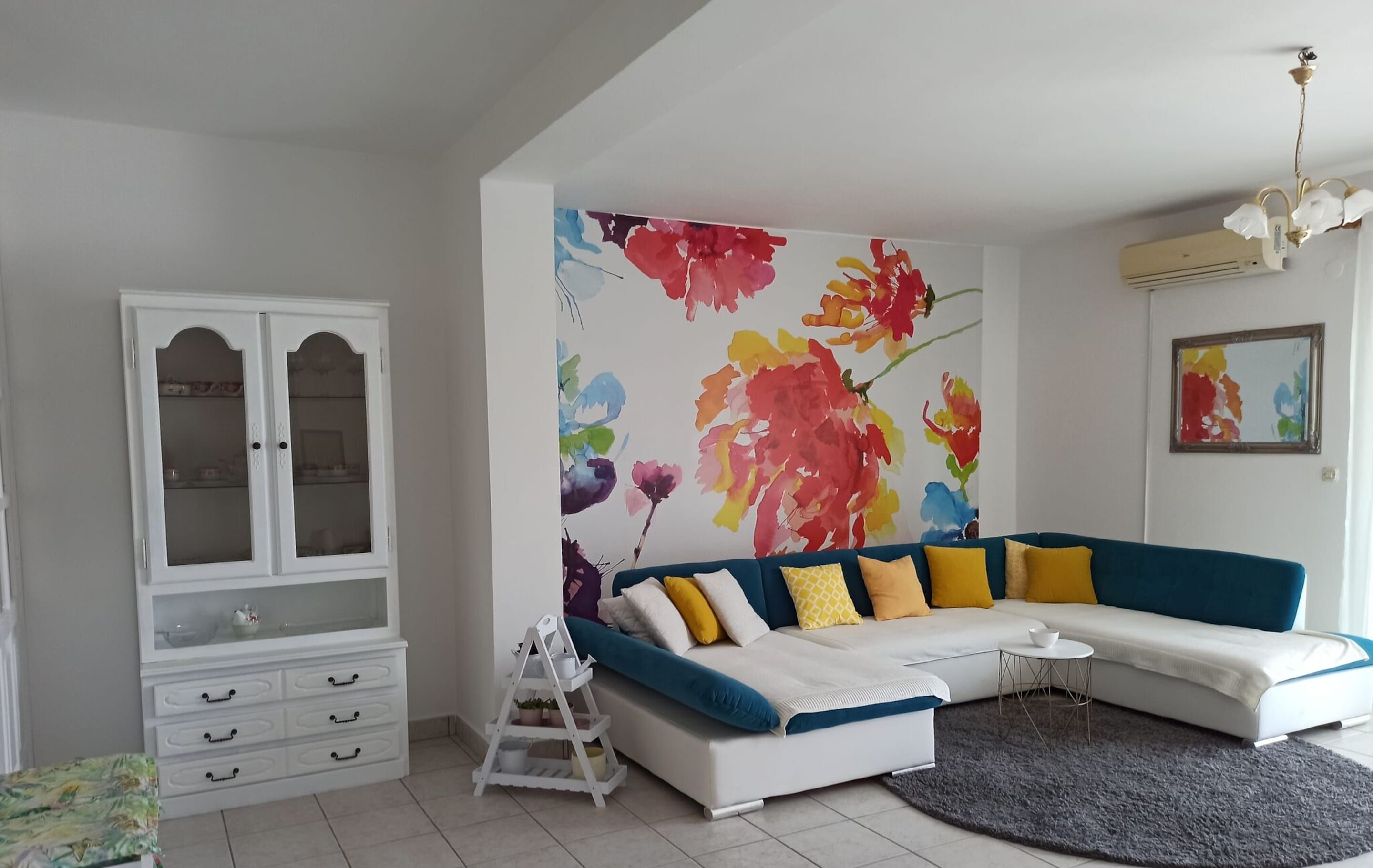 Una sala de estar con un sofá azul, cojines amarillos y blancos, un mueble blanco con puertas de cristal y un mural floral de colores.