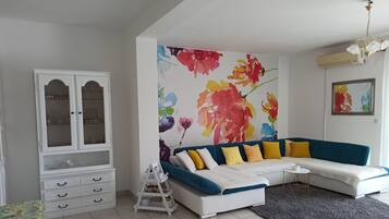 Una sala de estar con un sofá azul, cojines amarillos y blancos, un mueble blanco con puertas de cristal y un mural floral de colores.