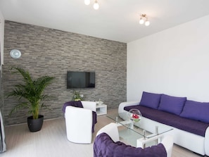 Uma sala de estar moderna com uma parede de tijolos cinza, uma televisão de tela plana montada acima de um sofá branco, uma mesa de centro de vidro e um vaso com planta.