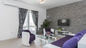 A modern living room with a stone wall, a flat-screen TV, a glass coffee table, and a sofa with purple cushions. - Apartment in Seget Donji Near Trogir Old Town (Seget)