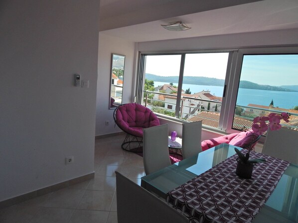 A balcony with a glass table, chairs, and a view of a lake and buildings. - Apartment in Okrug Gornji Near the Beach (Okrug)