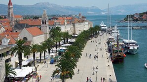 Une promenade en bord de mer bordée de palmiers, un bâtiment historique doté d’une tour et des bateaux amarrés dans la marina.