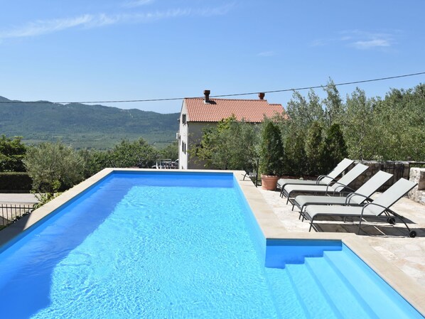 A rectangular swimming pool with clear blue water, surrounded by lounge chairs and potted plants, set against a backdrop of a house and mountains.