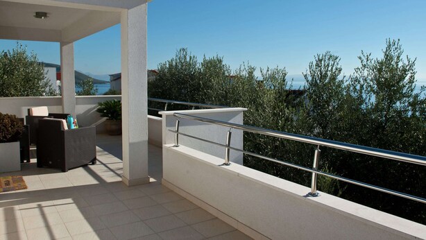 A modern outdoor balcony with a white railing, a seating area with a sofa and a table, and a view of the sea and greenery. - Holiday Home in Okrug Gornji Near the Sea (Okrug)