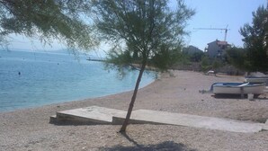 A pebble beach with a tree, a concrete pathway, and boats in the background.
